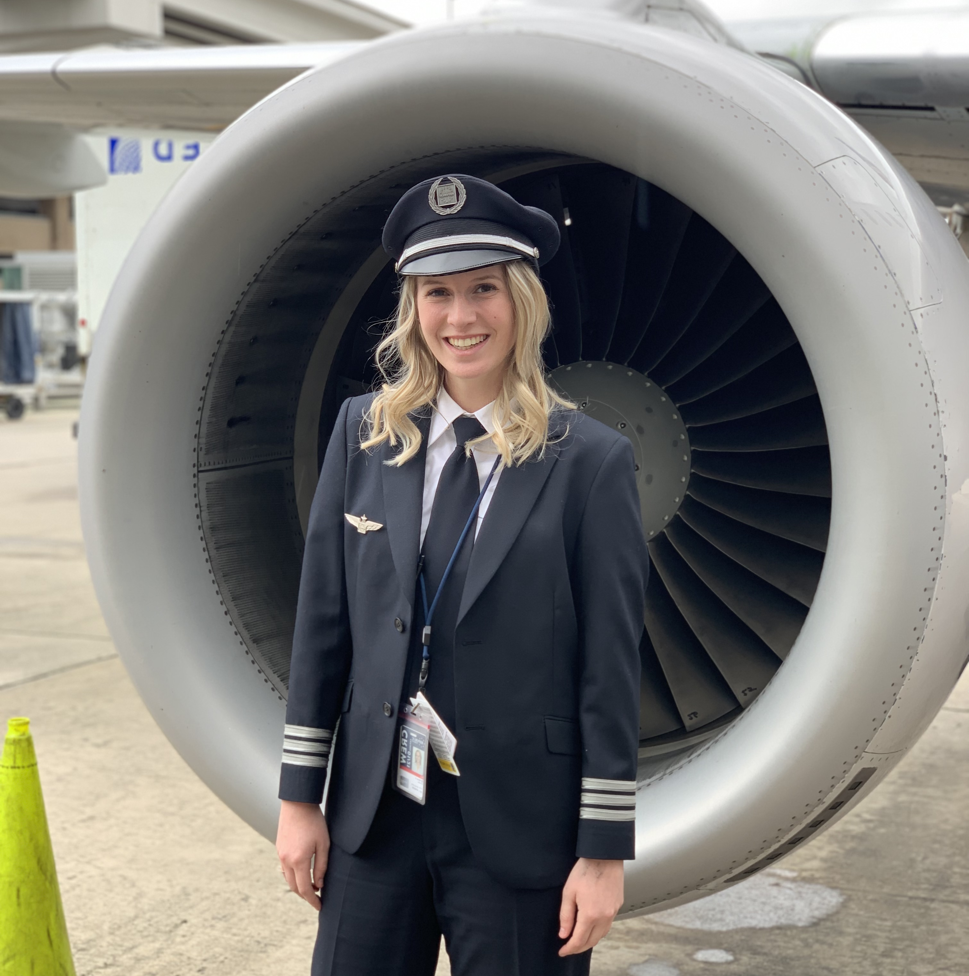 A smiling young blonde woman in a black and white pilot’s uniform and cap stands in front of an airplane’s silver-and-gray turbine engine.  