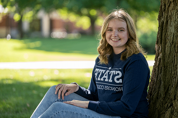 A female student with curly blonde hair sits with her back against a tree. She wears a dark blue STATE Honors College long-sleeved shirt and blue jeans. She is smiling and looking at the camera.